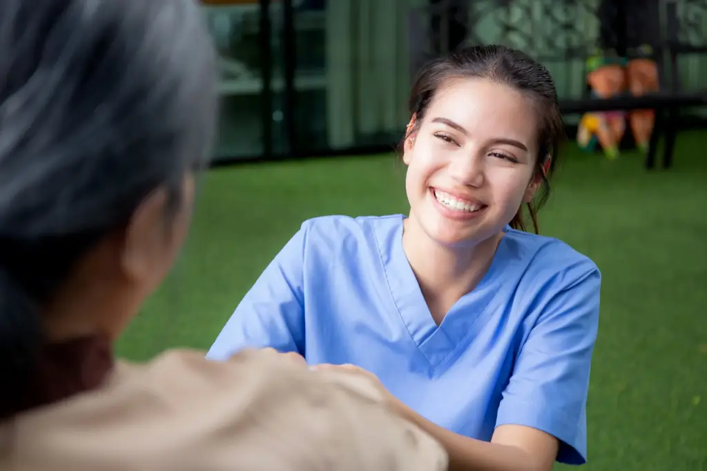 A smiling healthcare worker in blue scrubs interacts with an elderly person in an outdoor setting.